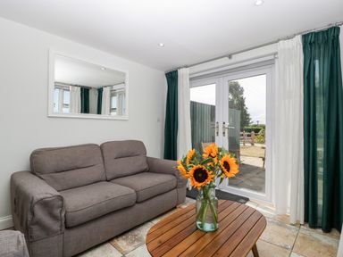 A living room with a grey sofa a wooden coffee table with sunflowers in a glass jar and glass doors with curtains at Somerset House in Othery near Middlezoy