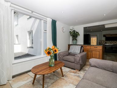 A living room with a wooden coffee table with sunflowers a grey armchair and sofa a plant on a cabinet and large window at Somerset House in Othery near Middlezoy