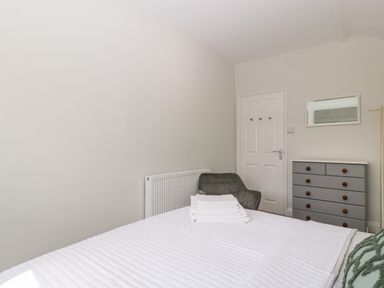 A bedroom with a white bedspread stacked with towels a grey armchair a grey chest of drawers a mirror and a door at Somerset House in Othery near Middlezoy