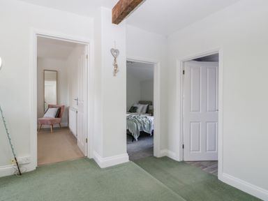 Hallway with green carpet and three doorways showing a pink chair with a cushion a bed with blankets and pillows and a closed white door at Somerset House in Othery near Middlezoy