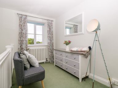 A small sitting area with a gray loveseat with cushions beside a window with floral curtains a white and gray dresser with a vase of flowers and folded blankets on top a floor lamp and a wall mirror at Somerset House in Othery near Middlezoy