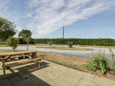 An outdoor space with a wooden picnic table on a paved area with plants and a curved gravel driveway surrounded by grass and hedges at Somerset House in Othery near Middlezoy