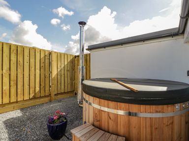 An outdoor hot tub with wooden exterior and a cover with a wooden scoop on top a wooden fence and a metal chimney pipe and a blue planter with flowers at Somerset House in Othery near Middlezoy