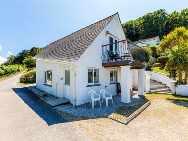 A white house with a balcony and plastic chairs outside near a curved staircase and trees at Baywatch Sands Polzeath