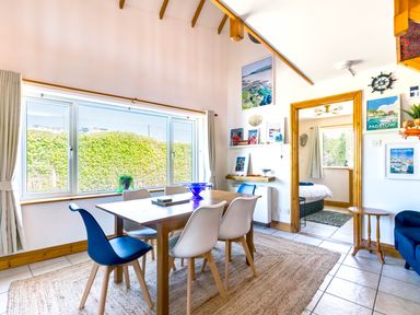 A dining area with a wooden table and six chairs next to a large window with hedges outside and view into a bedroom at Baywatch Sands in Polzeath