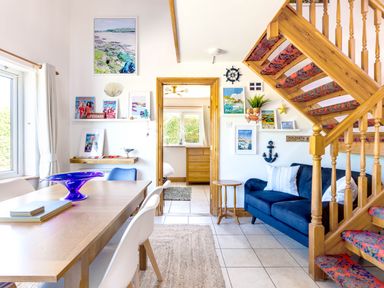 A dining area with a wooden table and chairs next to a blue sofa and wooden staircase at Baywatch Sands in Polzeath
