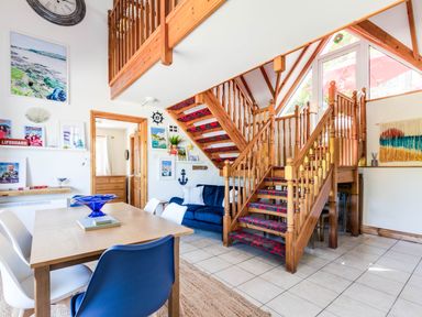 A dining area with wooden table and mixed chairs next to a staircase with wooden railings and red carpet at Baywatch Sands in Polzeath