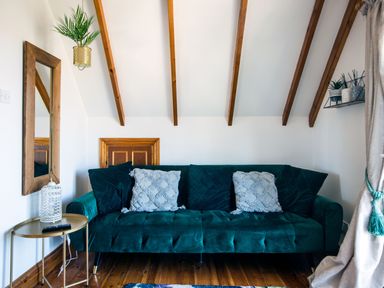 A green sofa with cushions next to a round side table with a lantern under a sloped ceiling with wooden beams at Baywatch Sands in Polzeath