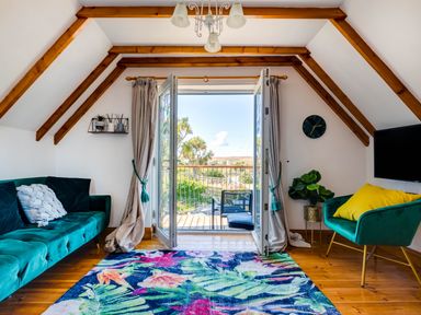 A room with wooden floor and ceiling beams with sofa armchair colorful rug and open double doors leading to a balcony at Baywatch Sands in Polzeath