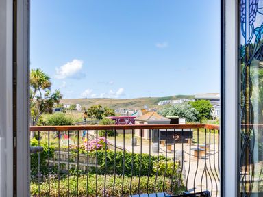 View through an open door showing a garden with flowers a fence and a playground with hills and buildings in the background at Baywatch Sands Polzeath