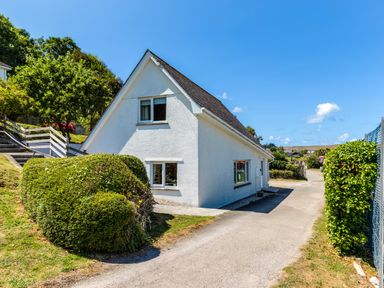 A white house with a sloped roof and three windows along a driveway with trimmed bushes and trees at Baywatch Sands in Polzeath