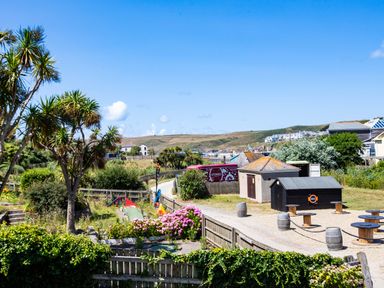 An outdoor area with palm trees flowers picnic tables and buildings with hills in the background at Baywatch Sands in Polzeath