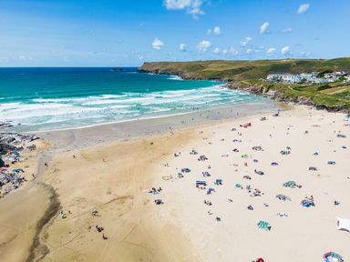 A sandy beach with people sunbathing and swimming with grassy hills and houses in the background at Baywatch Sands in Polzeath