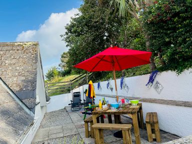 An outdoor patio with a wooden table and benches under a red umbrella with chairs and trees in the background at Baywatch Sands in Polzeath