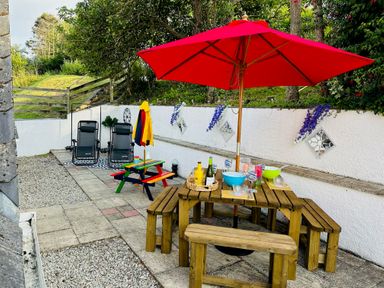 An outdoor patio with a wooden picnic table under a red umbrella and two black reclining chairs at Baywatch Sands in Polzeath