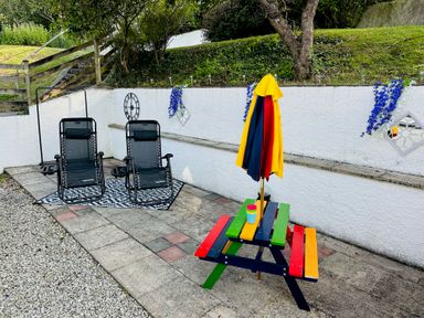 An outdoor patio with two black reclining chairs on a rug and a small colorful picnic table with a closed umbrella Baywatch Sands Polzeath