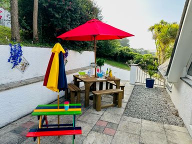An outdoor patio area with wooden tables and benches under a red umbrella and a small colorful bench with folded umbrellas at Baywatch Sands in Polzeath