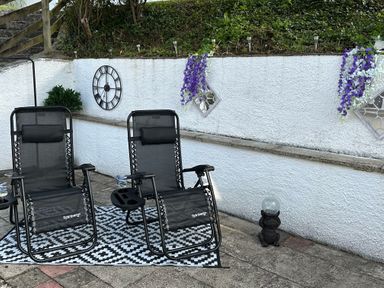Two black reclining chairs on a black and white patterned rug outside next to a white wall with purple flowers and a clock at Baywatch Sands in Polzeath