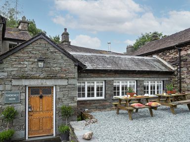 An outdoor patio area with wooden picnic tables and a stone building with a wooden door and multiple windows at Thorney How in Grasmere