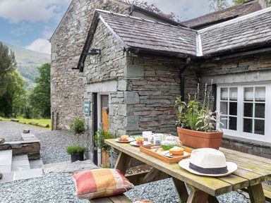 An outdoor wooden picnic table with a hat cushion plants and food outside a stone house at Thorney How in Grasmere