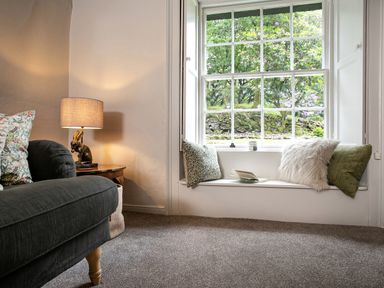 A corner of a living room with a dark sofa a wooden side table with a lamp and a window seat with cushions and a book at Thorney How in Grasmere