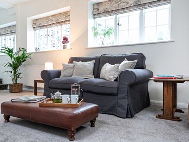 A living room with a gray sofa, brown ottoman, side table, plants, and windows at Thorney How in Grasmere