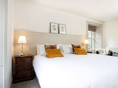 A bedroom with a large bed with white bedding and orange pillows two bedside tables with lamps and floral prints on the wall at Thorney How in Grasmere
