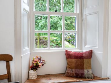A window with white wooden frames overlooking green trees a vase with flowers and a multicolored cushion on the wooden windowsill at Thorney How in Grasmere