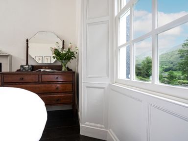 A wooden dresser with drawers a mirror and a vase of flowers beside a window showing a green landscape at Thorney How in Grasmere