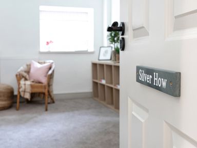 An open door with a silver how sign leading to a room with a wicker chair and cubby shelves at Thorney How in Grasmere