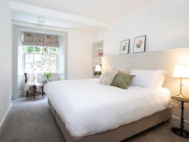 A bedroom with a large bed with pillows a chair with a blanket a small table with flowers and framed floral pictures at Thorney How in Grasmere