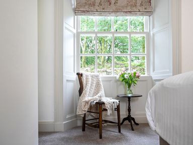 A wooden chair with a white knitted blanket next to a small round table with flowers by a window at Thorney How in Grasmere