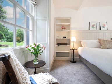 A bedroom with a bed pillows a bedside lamp shelves with decor a chair with a blanket next to a window with a vase of flowers at Thorney How in Grasmere