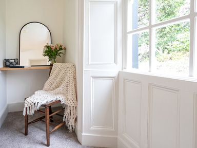A wooden chair with a knitted blanket beside a wall-mounted shelf with a mirror flowers and books at Thorney How in Grasmere
