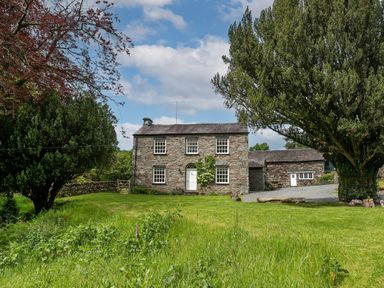 A stone house with a white door and five windows surrounded by green grass and trees at Thorney How in Grasmere