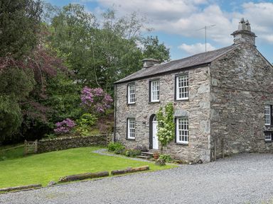 A stone house with white windows and a door surrounded by a green lawn and trees at Thorney How in Grasmere