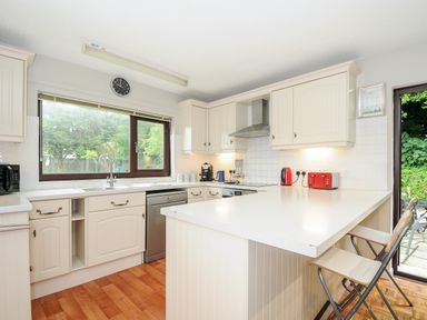 A kitchen with white cabinets wooden floor a large window and two stools at Spring Lodge in Eastcott near Kilkhampton