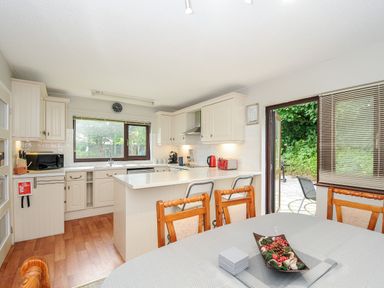 A kitchen with white cabinets a dining table with wooden chairs and a window showing greenery at Spring Lodge in Eastcott near Kilkhampton