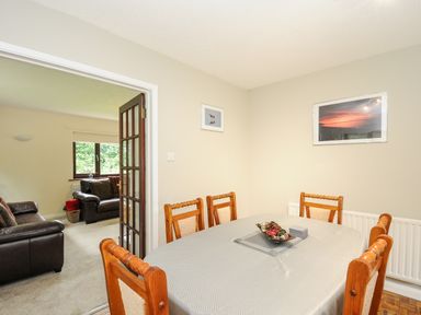 A dining room with a table covered by a tablecloth and six chairs next to an open door to a living room with two leather sofas at Spring Lodge in Eastcott near Kilkhampton
