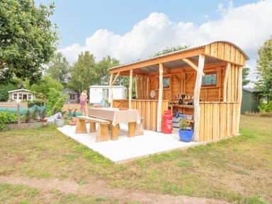 An outdoor wooden shelter with a picnic table and benches in a garden with trees and sheds at Willow Cottage in Hickling near Catfield