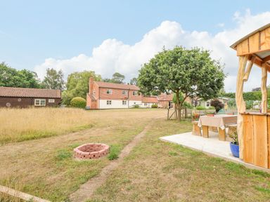 A backyard with a brick fire pit a wooden picnic table beneath a pergola a tree and a brick house in the background at Willow Cottage in Hickling near Catfield