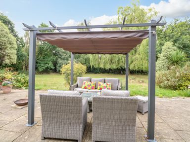 A patio with outdoor wicker furniture under a pergola with a garden in the background at Willow Cottage in Hickling near Catfield