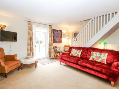 A living room with a red sofa and floral cushions an orange armchair and wooden chairs near a staircase at Willow Cottage in Hickling near Catfield