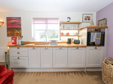 A kitchen with white cabinets wooden countertops a window with a pink blind and shelves with kitchen items at Willow Cottage in Hickling near Catfield