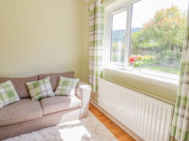 A living room with a beige sofa with green and white plaid cushions by a window with green plaid curtains and a vase of flowers on the windowsill at Cam O'r Fechan Annexe in Llangollen