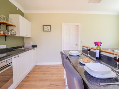 A kitchen with white cabinets wooden shelves glasses a countertop with plates bread wine glasses and chairs at Cam O'r Fechan Annexe in Llangollen