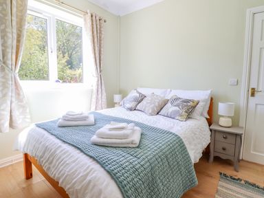 A bedroom with a bed pillows and folded towels on a blanket beside a window with curtains and a bedside table with lamp at Cam O'r Fechan Annexe in Llangollen