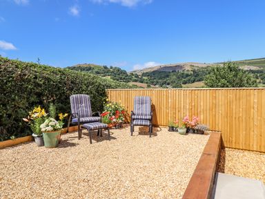 An outdoor patio area with two striped chairs and potted flowers surrounded by a wooden fence and hedge at Cam O'r Fechan Annexe in Llangollen