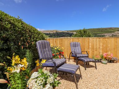 A garden with two striped chairs and footrests surrounded by flowers and a wooden fence at Cam O'r Fechan Annexe in Llangollen