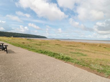 A paved walkway with benches beside a grassy area and a beach at Bluebell Lodge Meadow view lodges in Brean